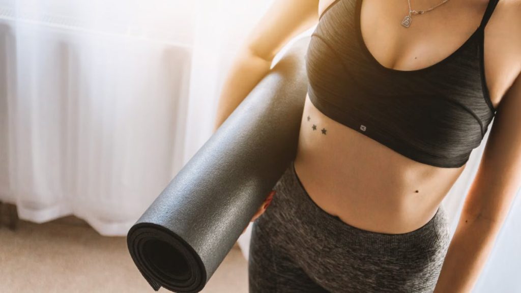 Fit woman in active wear holding yoga mat indoors, ready for workout.